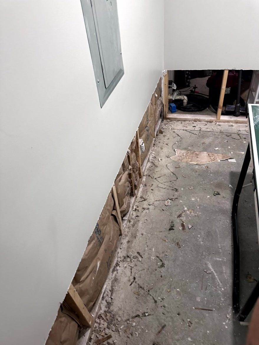 A room corner with damaged drywall exposing wood studs and brown insulation, near a floor-level electrical panel.
