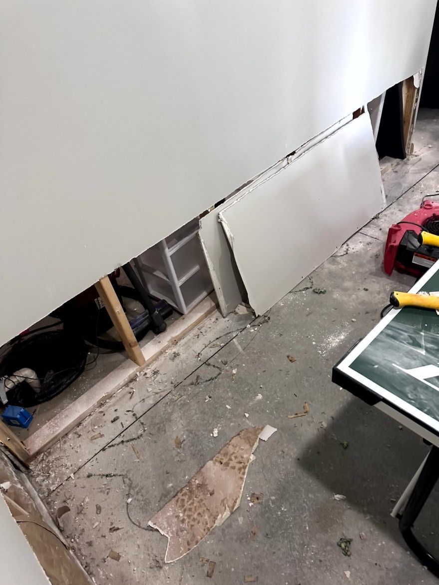 A room under renovation with a section of wall drywall removed, revealing the wooden frame and floor debris below.