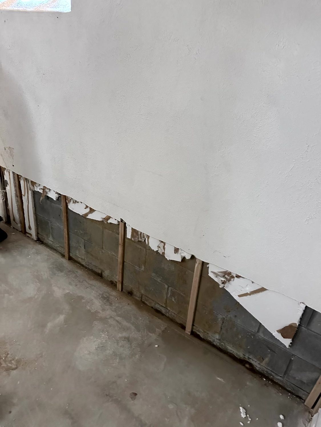 Damaged, unfinished wall showing drywall torn away to reveal wooden studs and gray concrete blocks underneath.