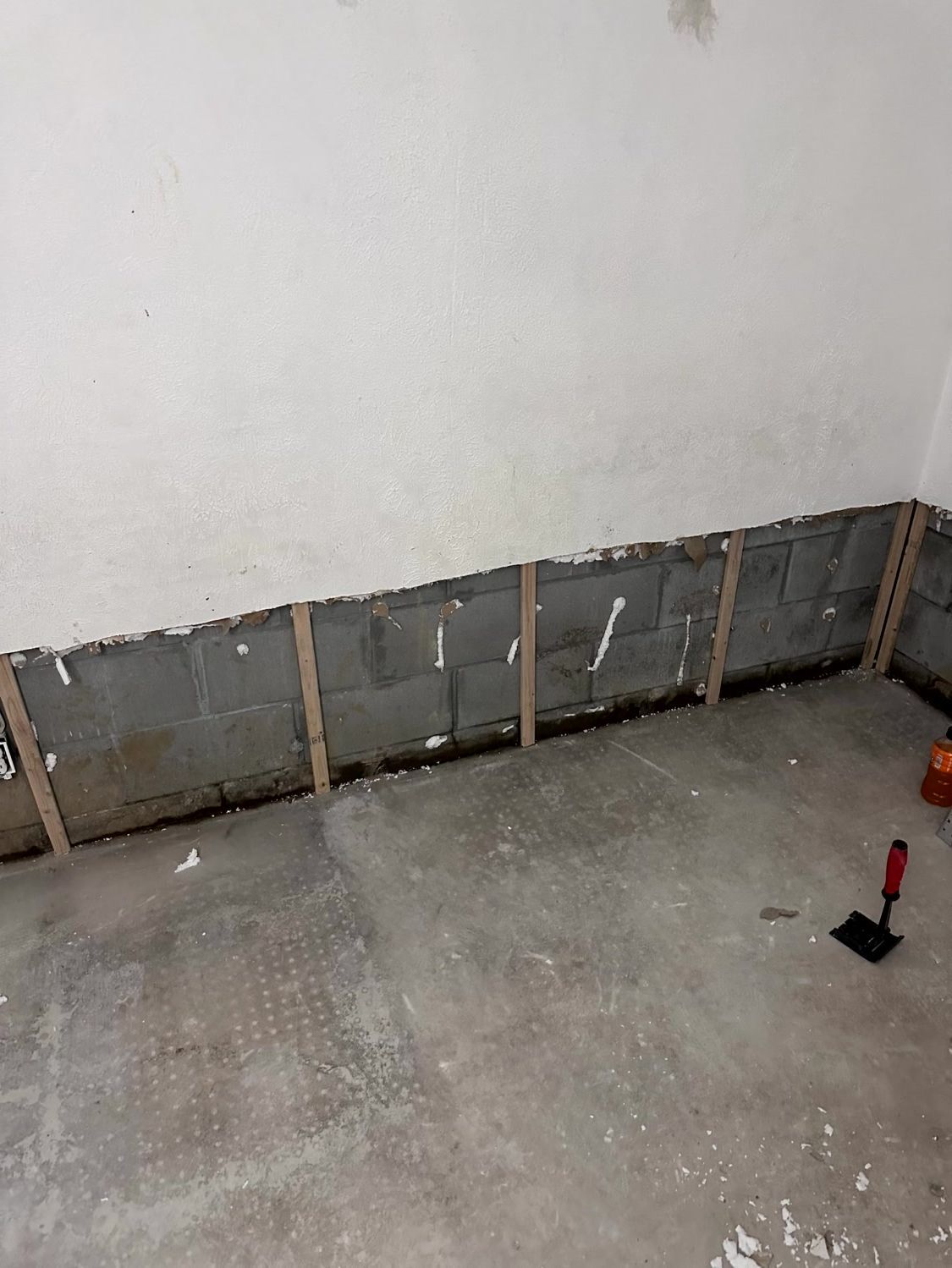 A basement wall with exposed concrete blocks and vertical wooden furring strips above a concrete floor.