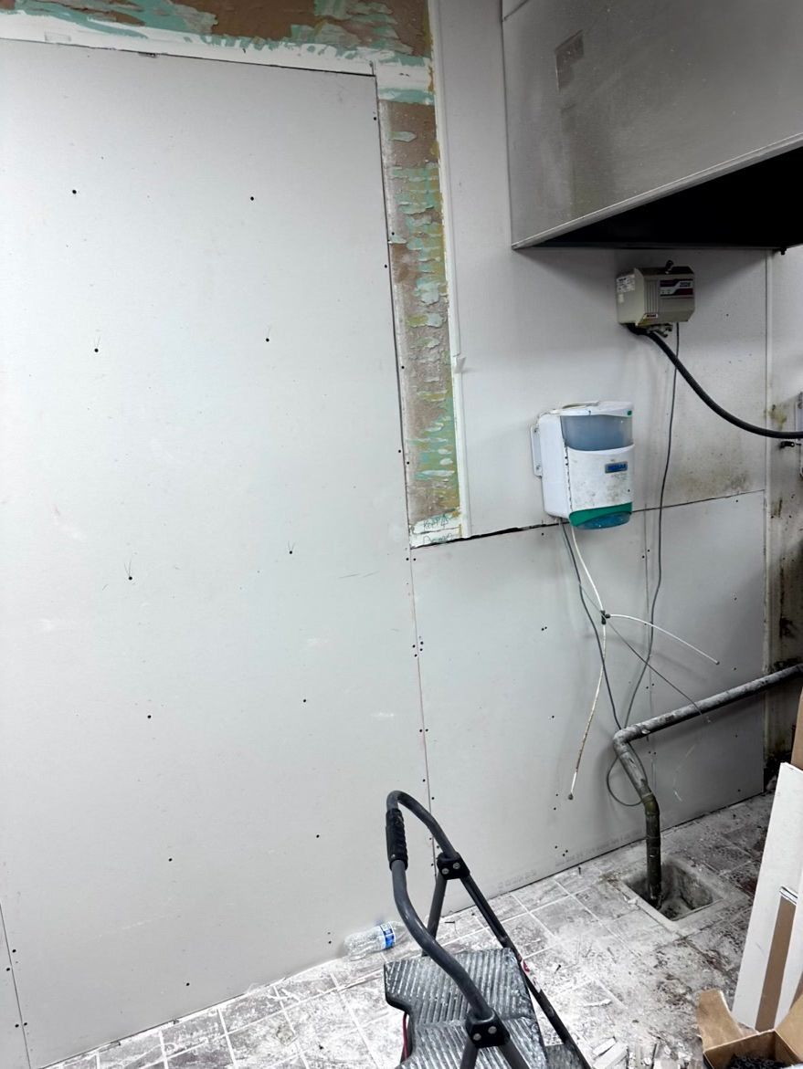 A partially finished wall with new white drywall panels, a wall-mounted dispenser, exposed wiring, and construction debris.