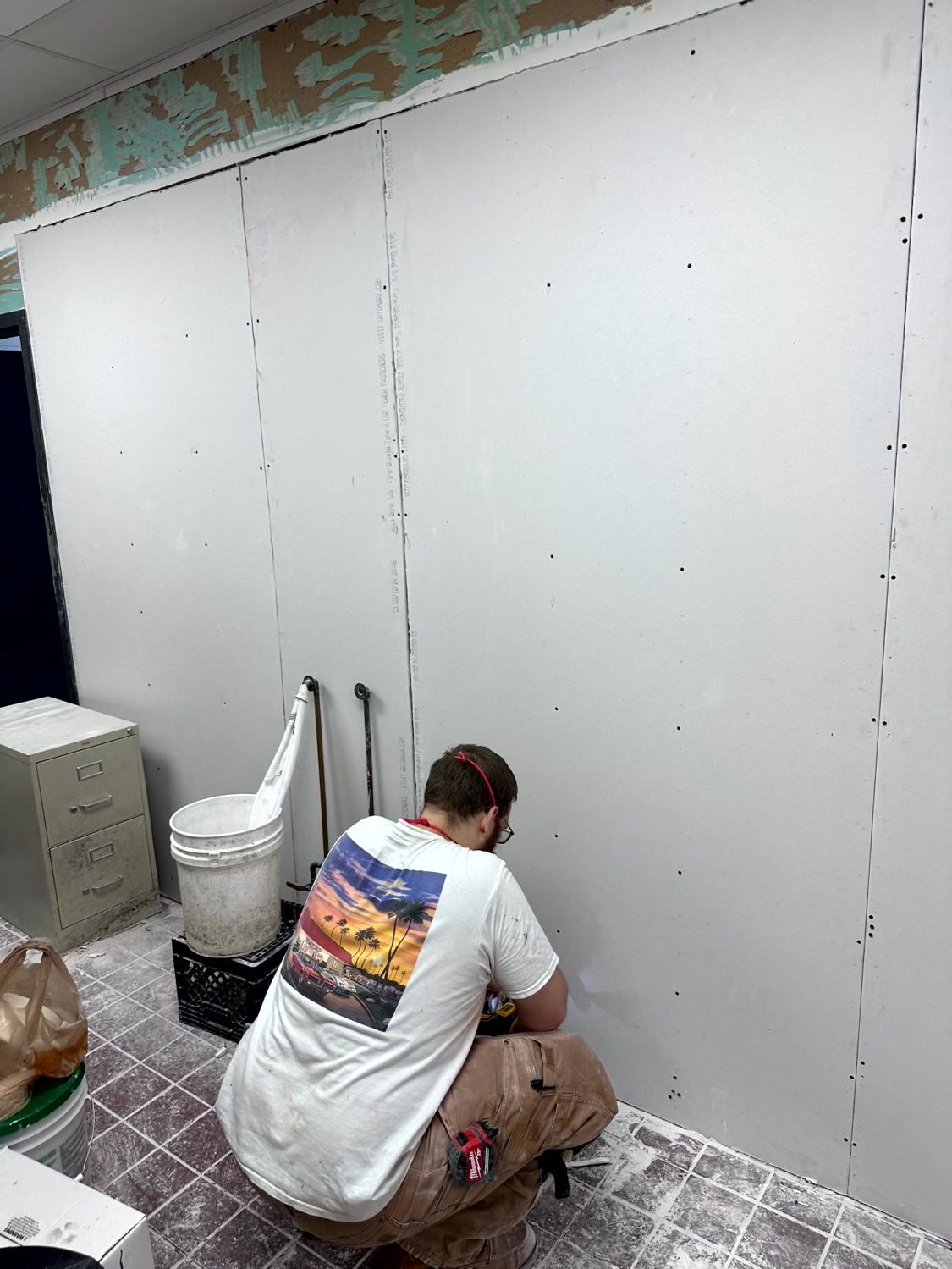 A person in a white t-shirt and tan pants kneels on a dusty floor, installing drywall panels on a large interior wall.