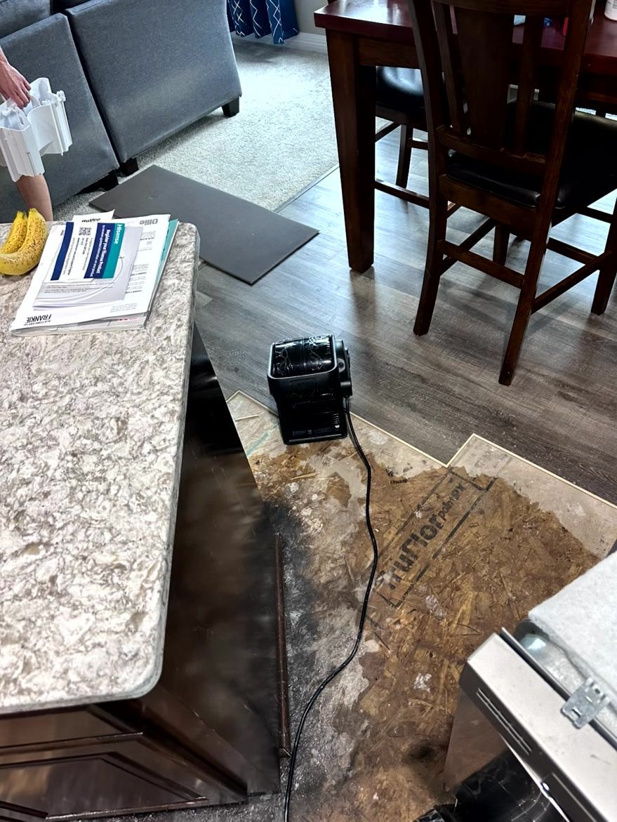 A floor fan on exposed plywood subflooring in a room with a kitchen island and dining furniture.