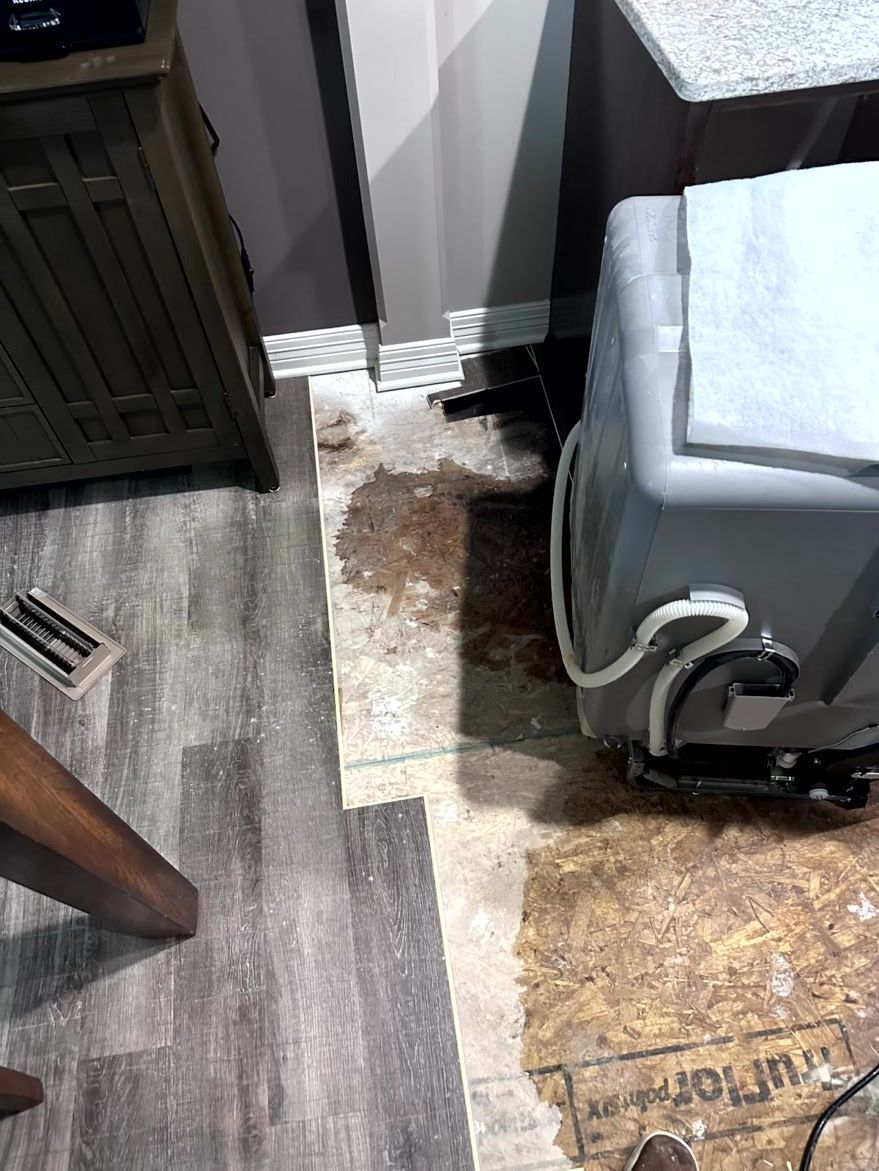 A kitchen floor with exposed subflooring and a dark, damp stain near a dishwasher and a cabinet base.
