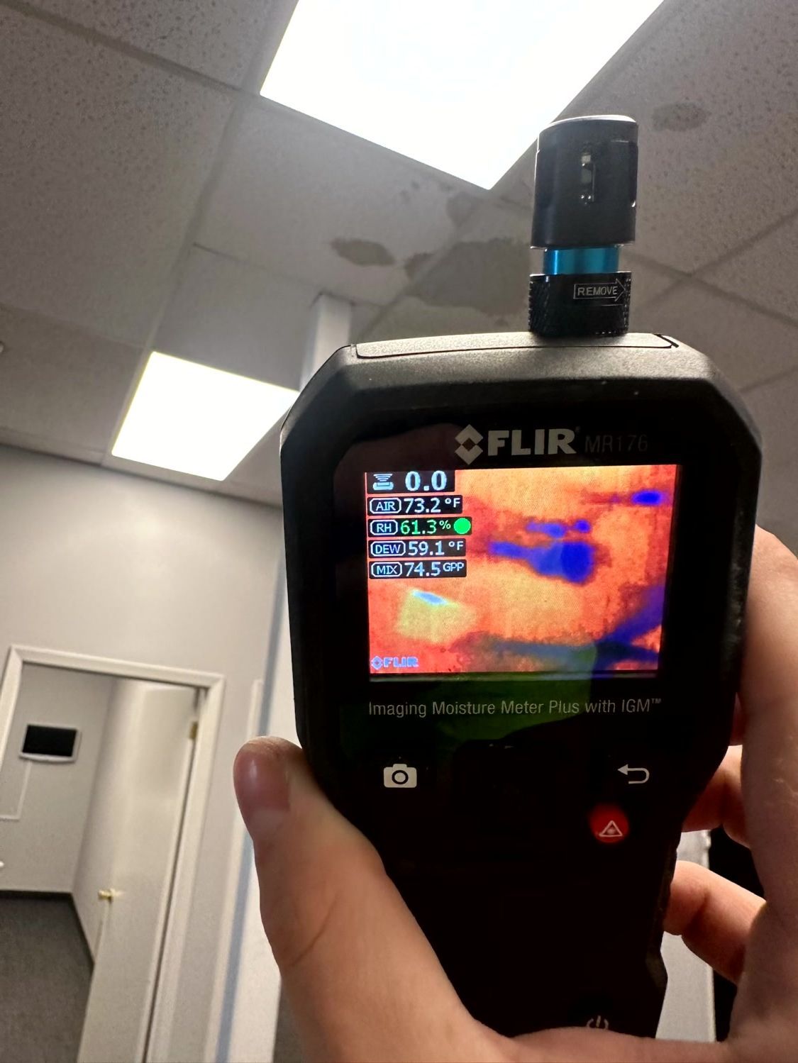 Hand holding a FLIR thermal imaging camera pointing at a ceiling with water damage, displaying a colorful heat map.