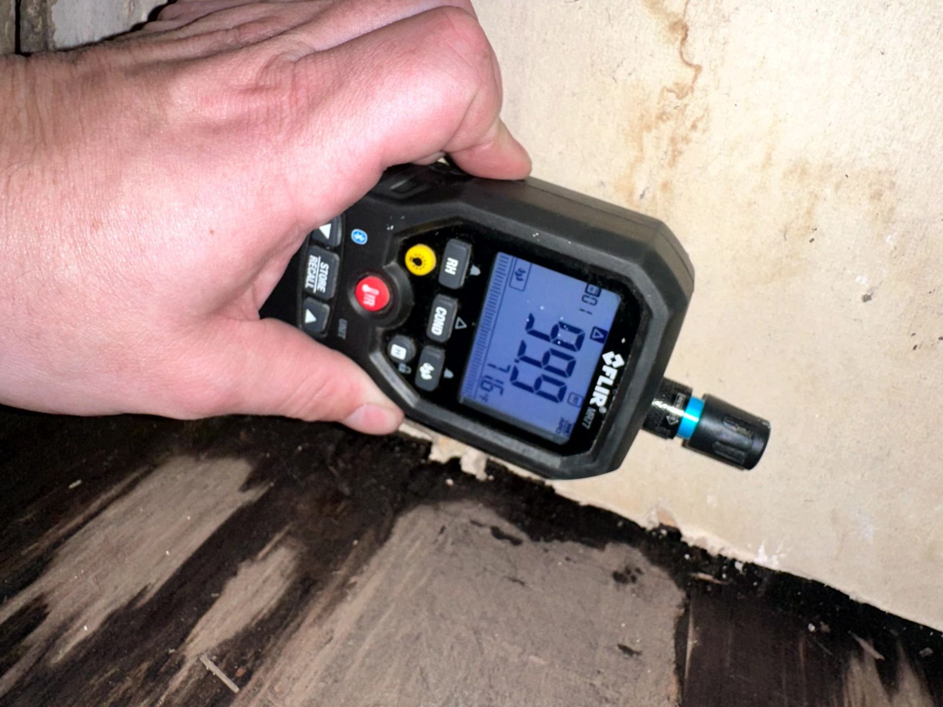 A hand holds a moisture meter against a water-damaged wall above dark, warped floorboards; the meter displays 99.9.