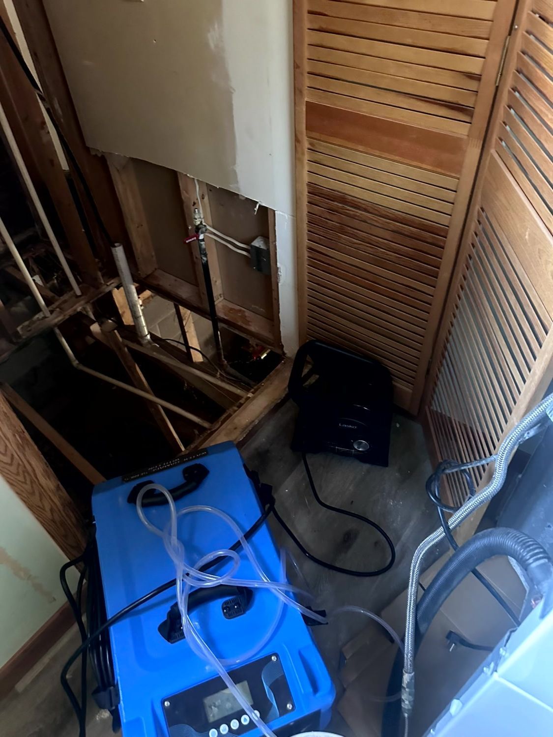 A blue commercial dehumidifier and an air mover sit on a floor next to a wall with a hole exposing interior framing.