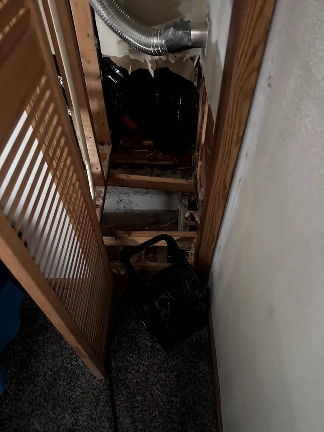 An open closet wall revealing a dark, recessed crawl space with wooden framing, metal ductwork, and debris.