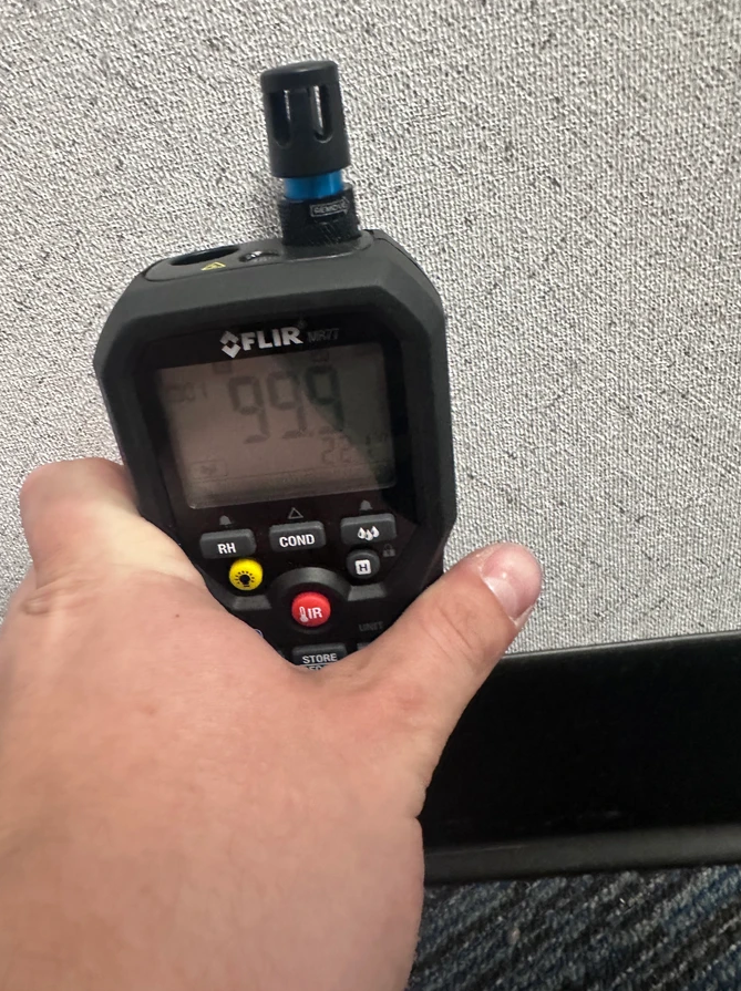 A hand holds a black FLIR moisture meter against a grey office cubicle wall, displaying a reading of 99.9.