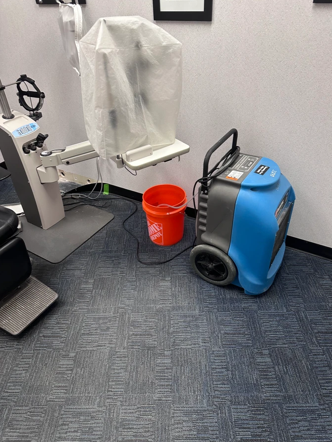 A blue dehumidifier sits next to an orange bucket in a room with a covered medical exam chair.