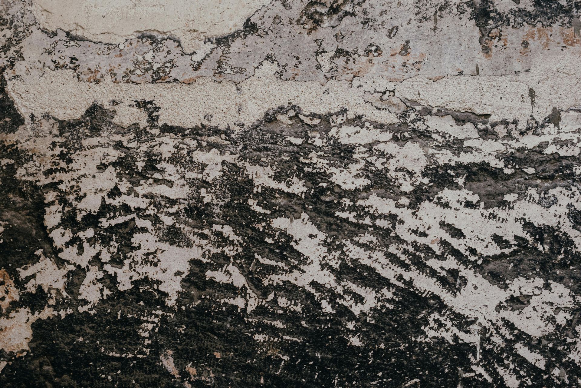 A close-up of a weathered, textured wall with patches of peeling white paint over dark, uneven concrete.