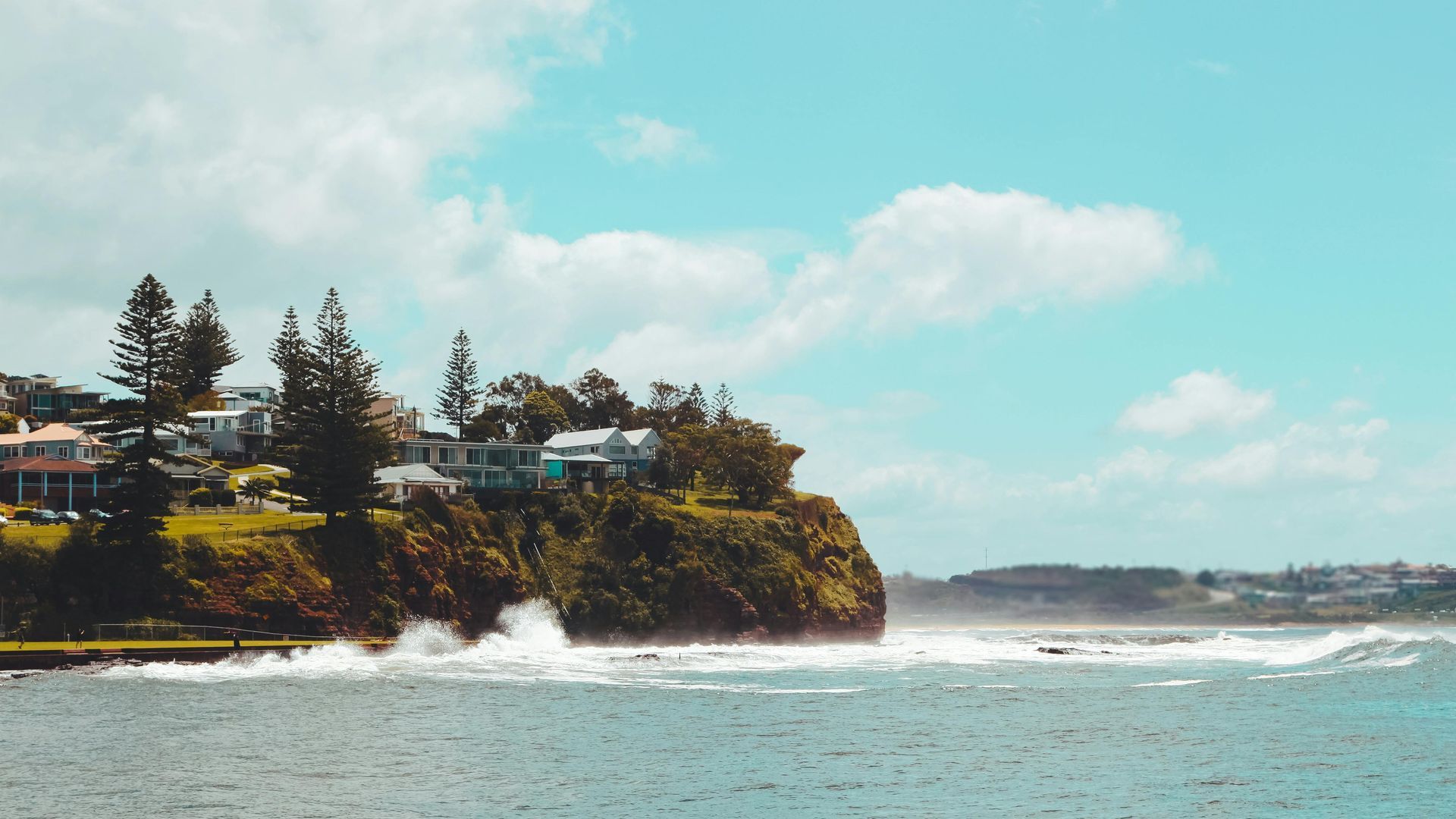 Cliffs with seaside houses and tall trees above crashing waves under a blue sky