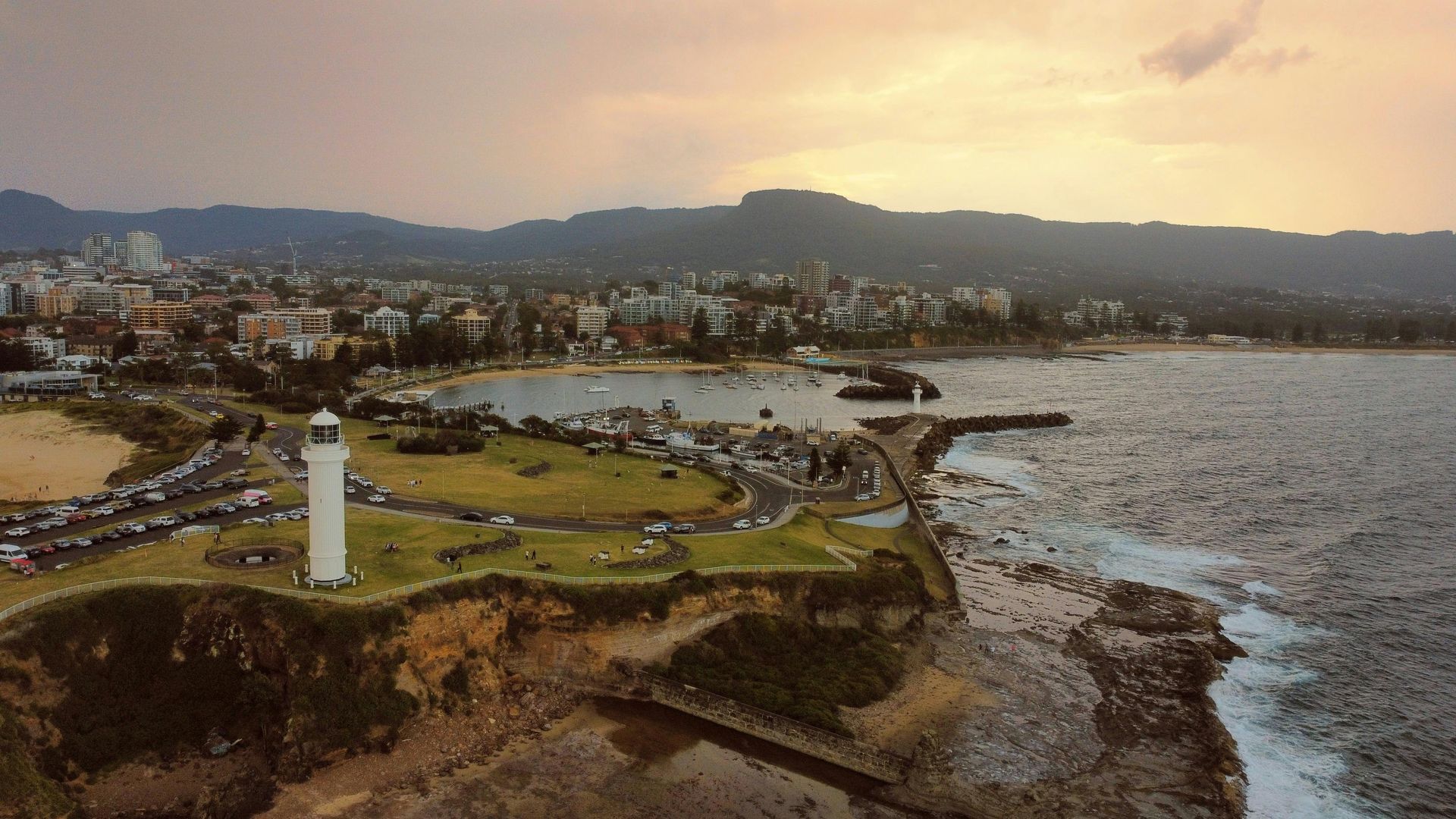 Coastal lighthouse at sunset beside rocky shore, with harbor, city, and mountains in the distance