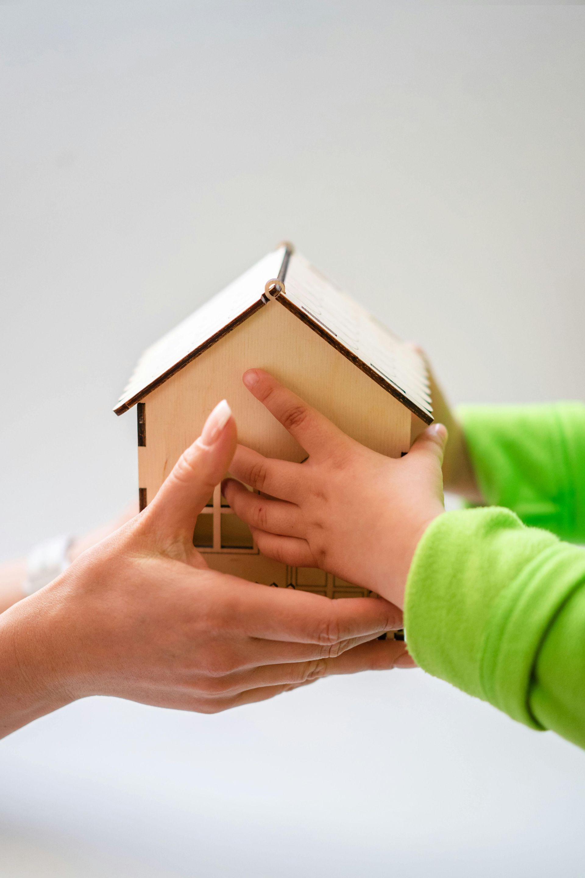 An adult's hands and a child's hands in a green sleeve hold a small, wooden model house together against a white background.
