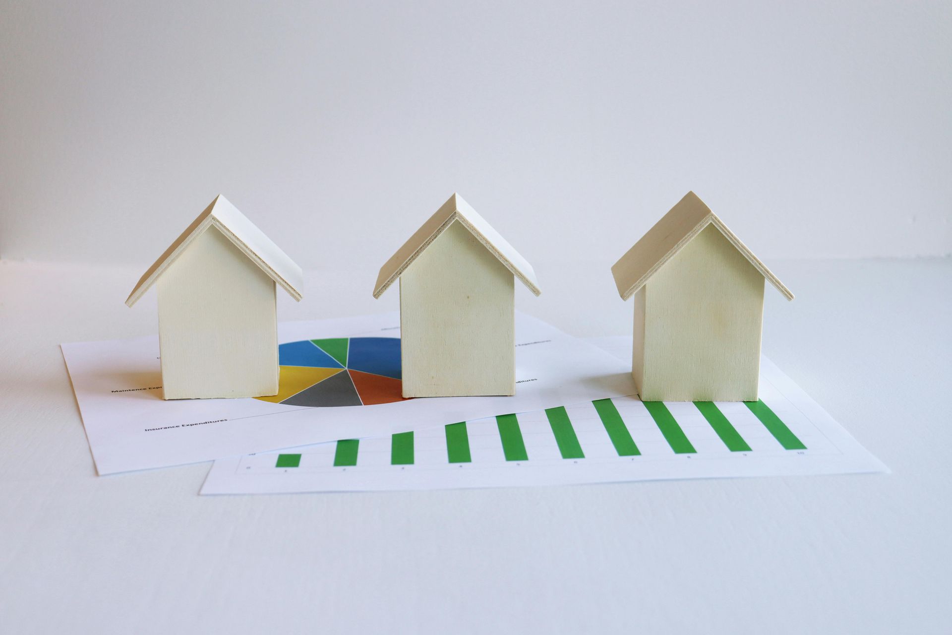 Three small white paper houses on a white surface with green and blue chart-like bars in front