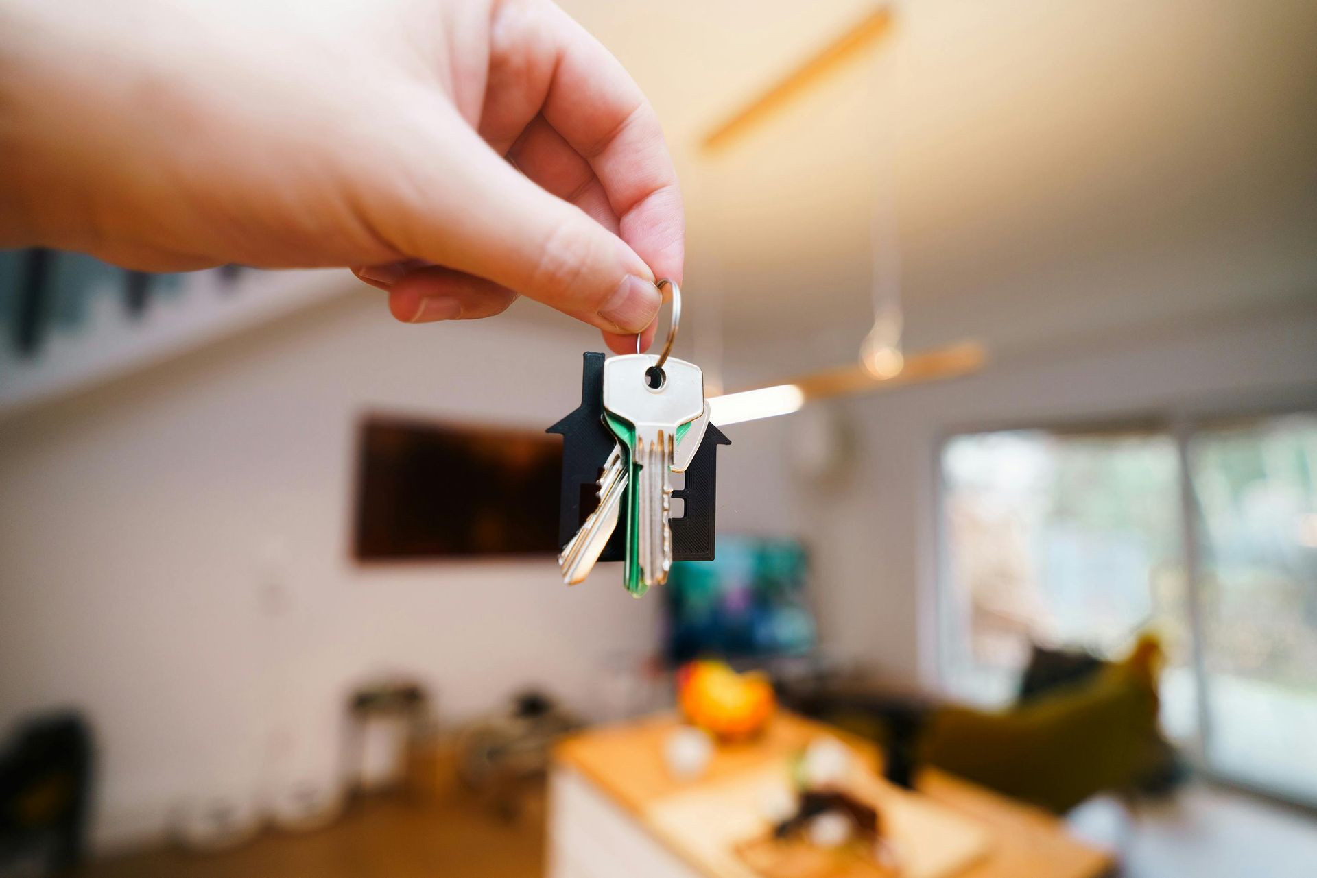 Hand holding a small keychain in a bright living room with a blurred couch and window background