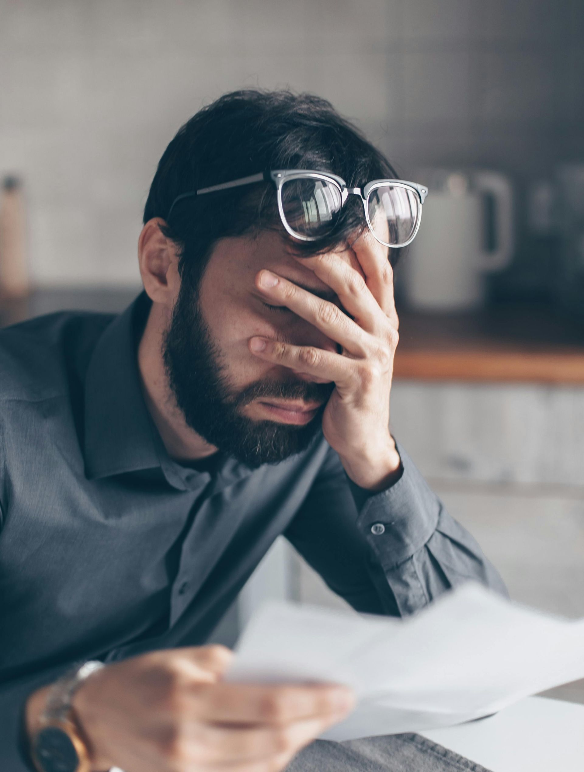A person in a gray shirt sits at a desk with papers, covering their face with one hand while wearing glasses on their head.