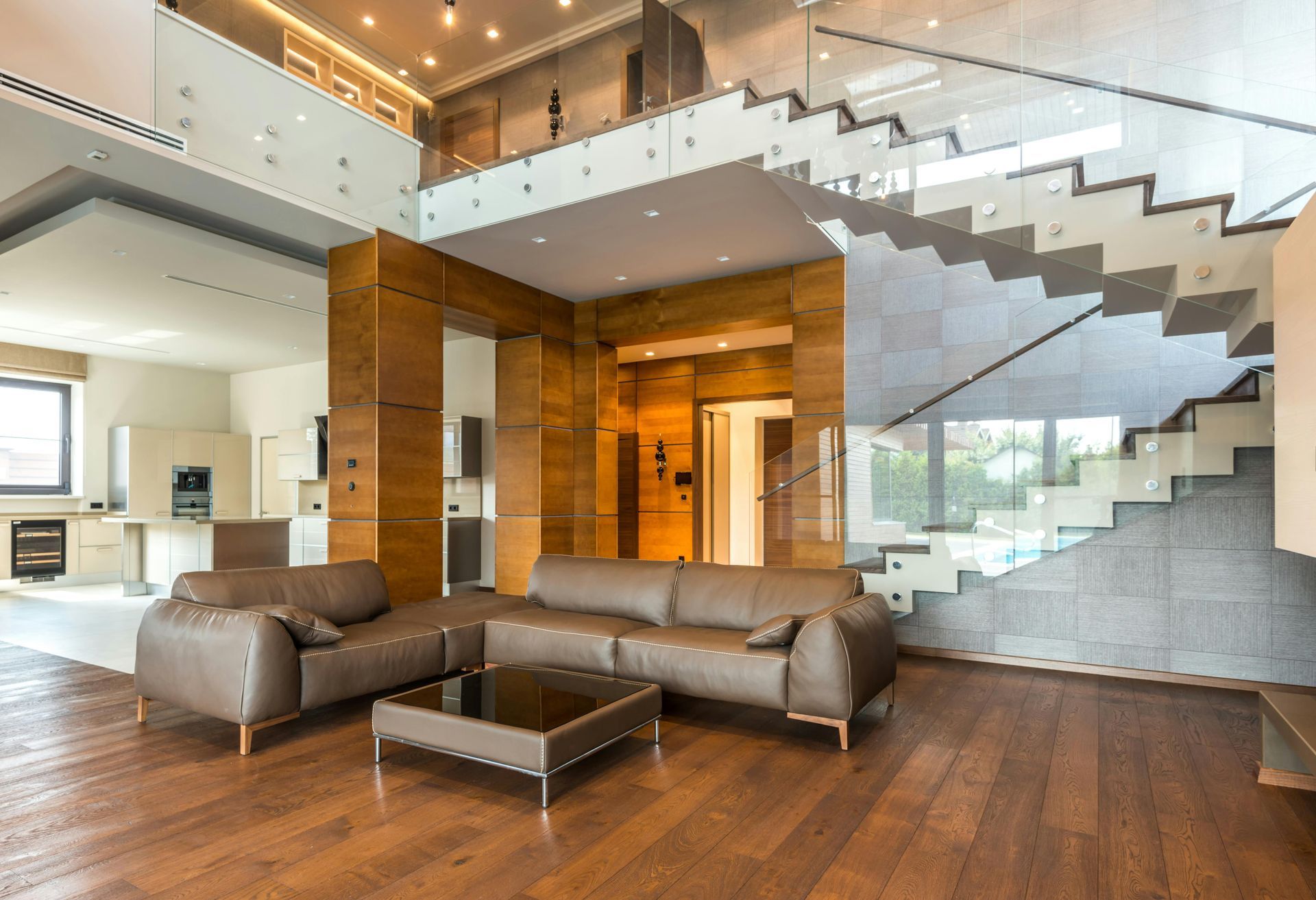 Modern living room with a brown leather sectional, glass coffee table, and an open staircase against a stone wall.