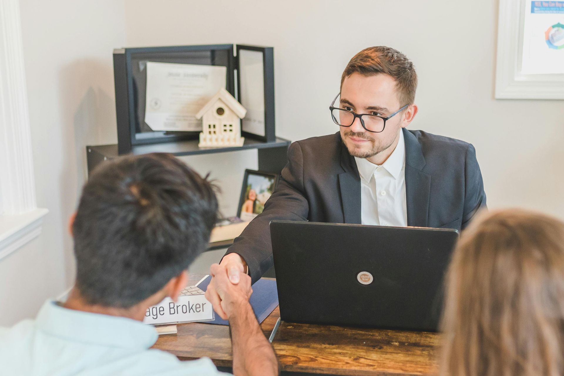 A professional in a suit shakes hands with a client across a desk with a laptop, with a small model house in the background.