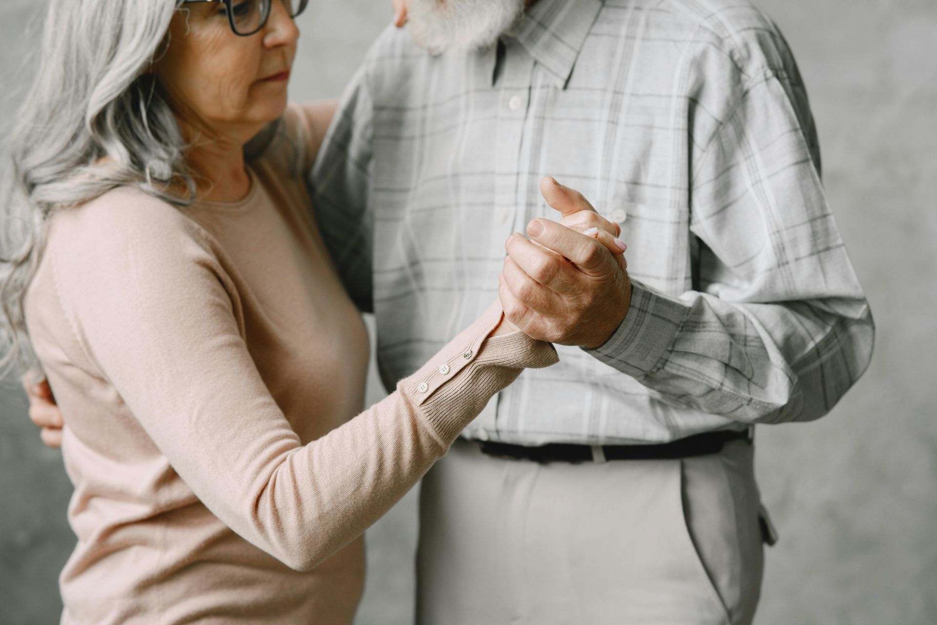 A retired couple dancing, holding hands in a slow, graceful pose against a plain gray wall.