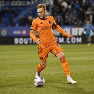 A man in an orange jersey is dribbling a soccer ball on a field.