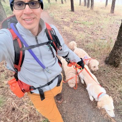 Man hiking with three dogs on a trail in a forest. He is wearing glasses, a hat, and carrying a backpack.