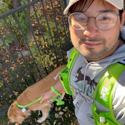 Man in glasses and cap walks golden retriever wearing a green harness and leash.