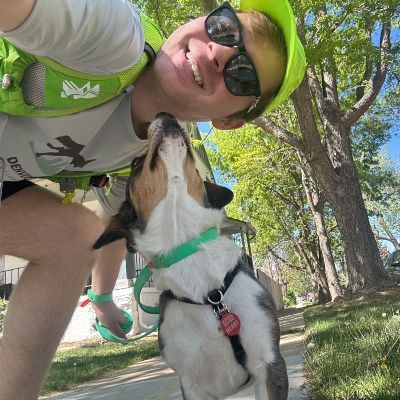 Scott in neon hat and sunglasses kneels beside a dog in a park, both sharing a close nuzzle.