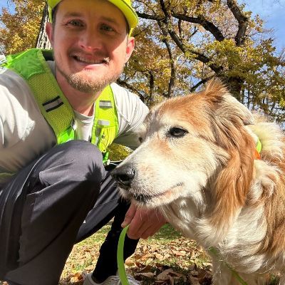 Man in safety gear kneels next to a fluffy dog, both in a park setting.