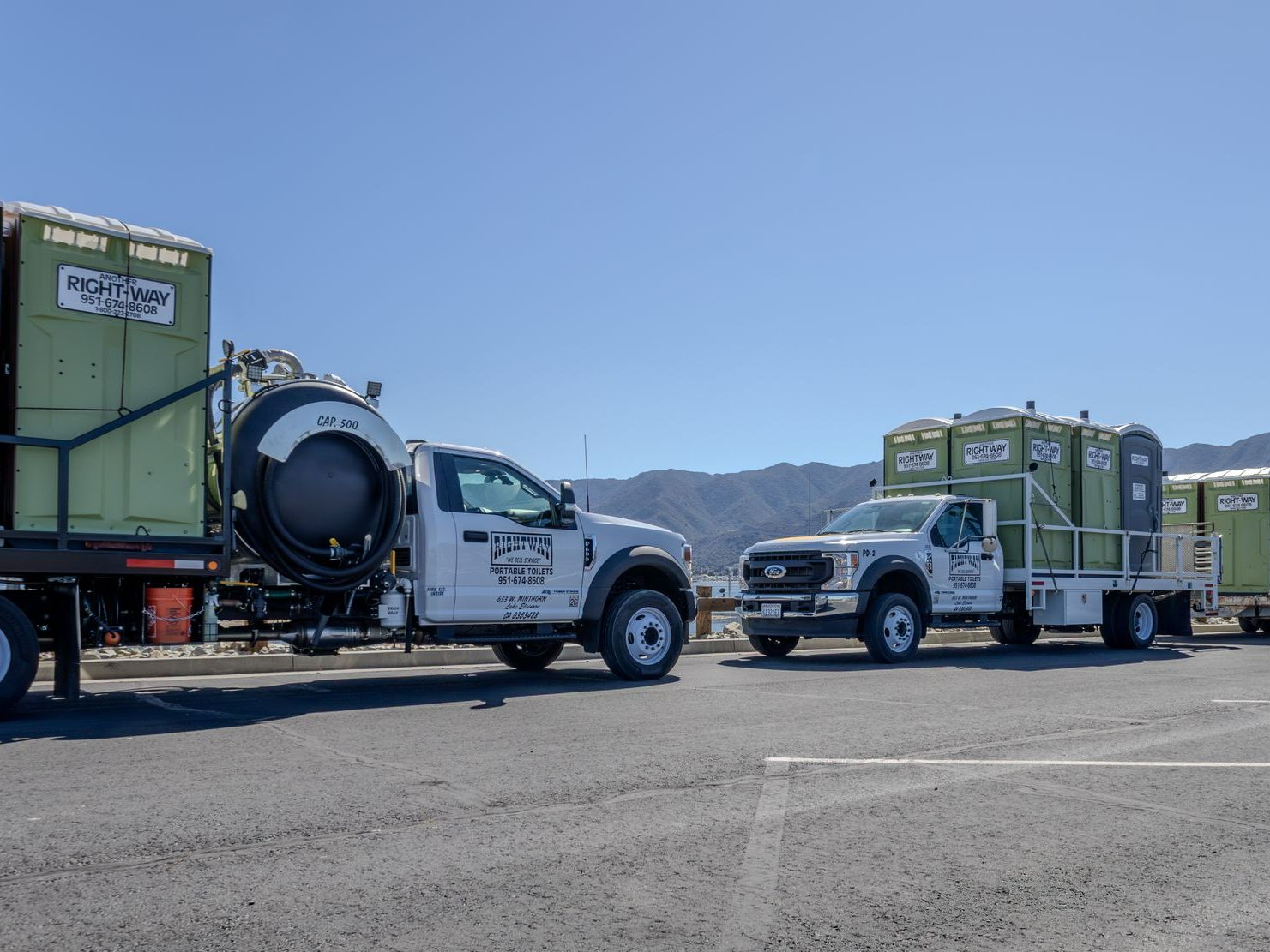 Two white trucks with portable toilets, parked in an outdoor lot. The trucks bear company signage.