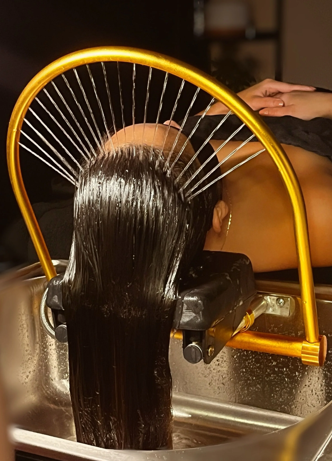 Person having hair rinsed in a salon sink with an arched water-spraying device. Dark hair, gold frame, and stainless steel sink.
