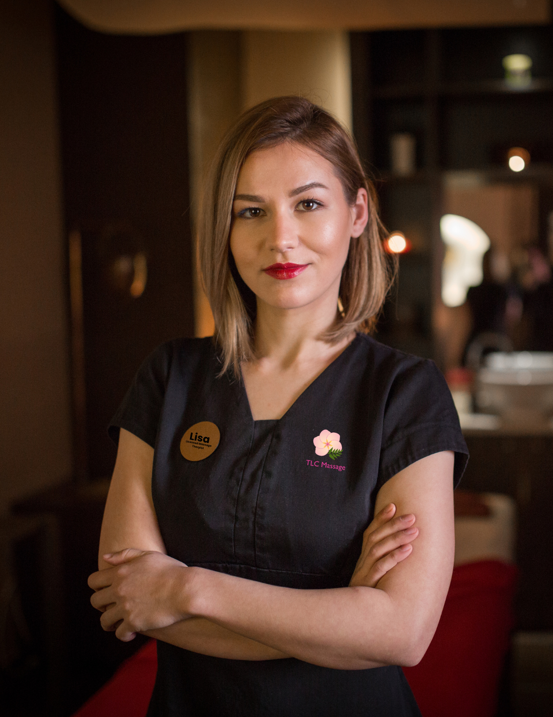 A female licensed massage therapist in a black shirt is standing with her arms crossed in a massage parlor