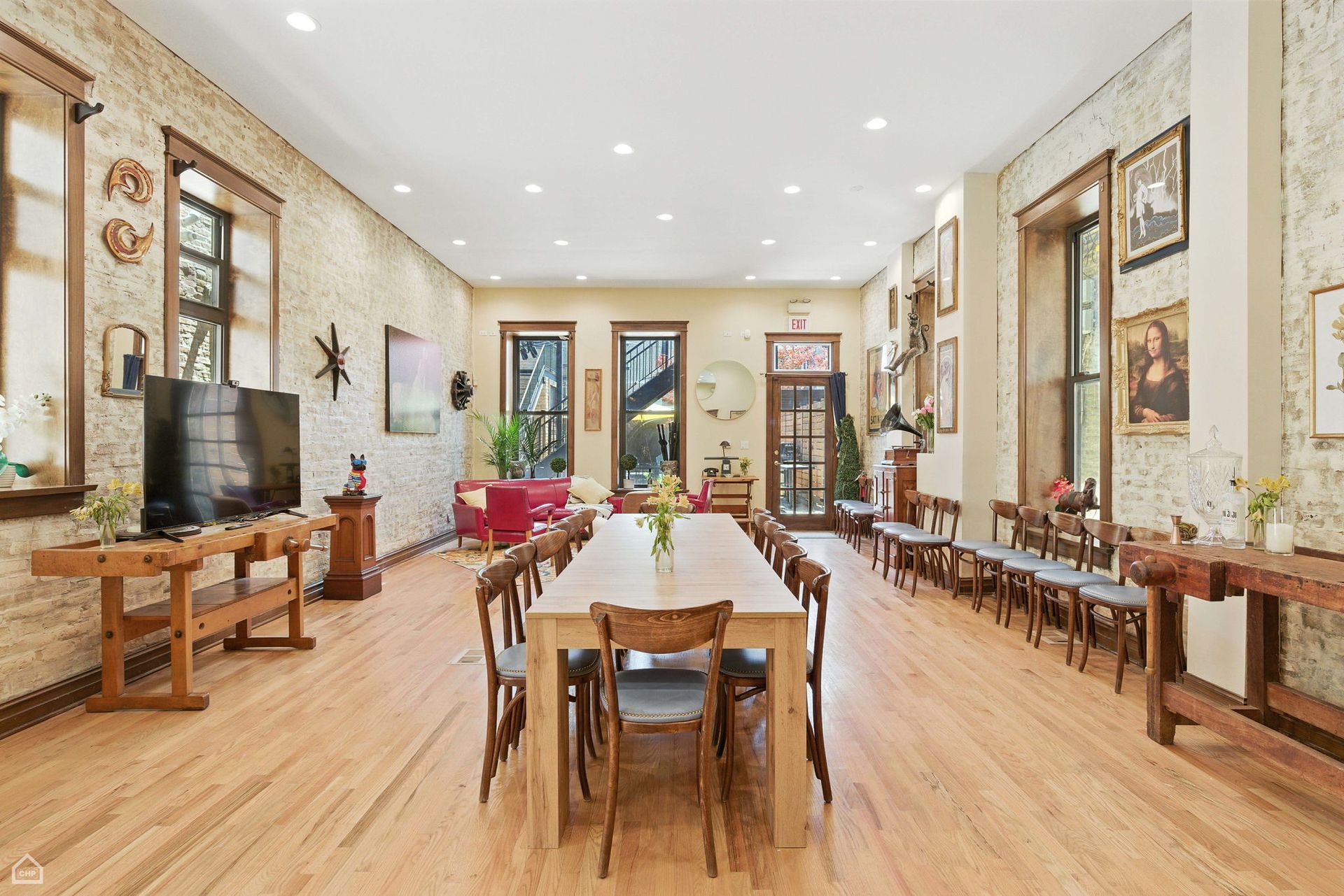 Large, light-filled dining room with long table, chairs, wood floors, exposed brick walls, windows, and artwork.