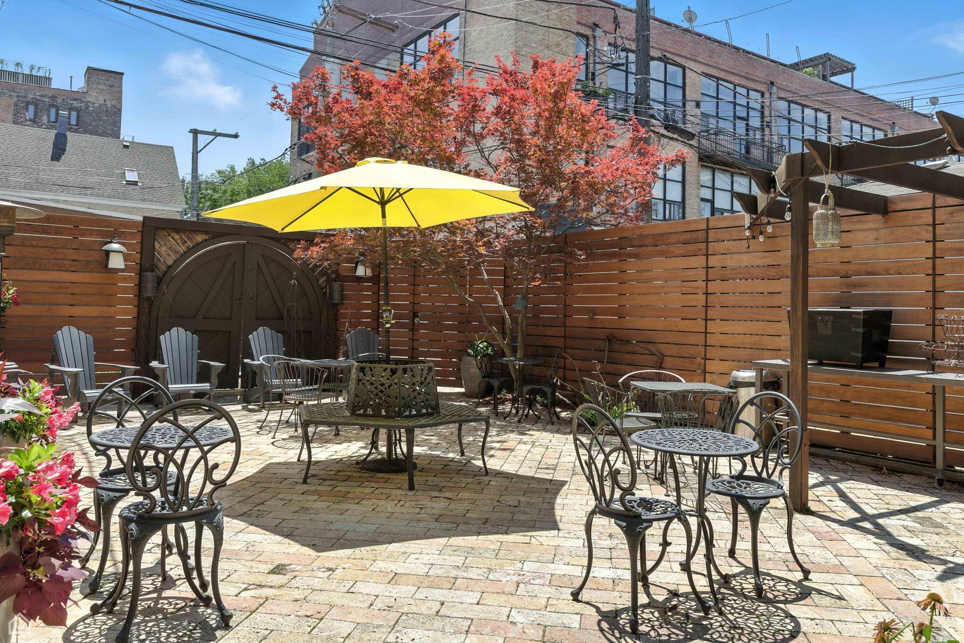 Outdoor patio with iron tables, chairs, and yellow umbrella, enclosed by wooden fence and brick ground.