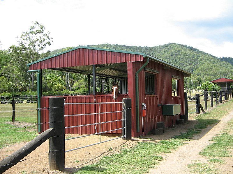 A Red Building With A Green Roof Is In A Field With Mountains In The Background — Austeel Sheds in Bowen, QLD