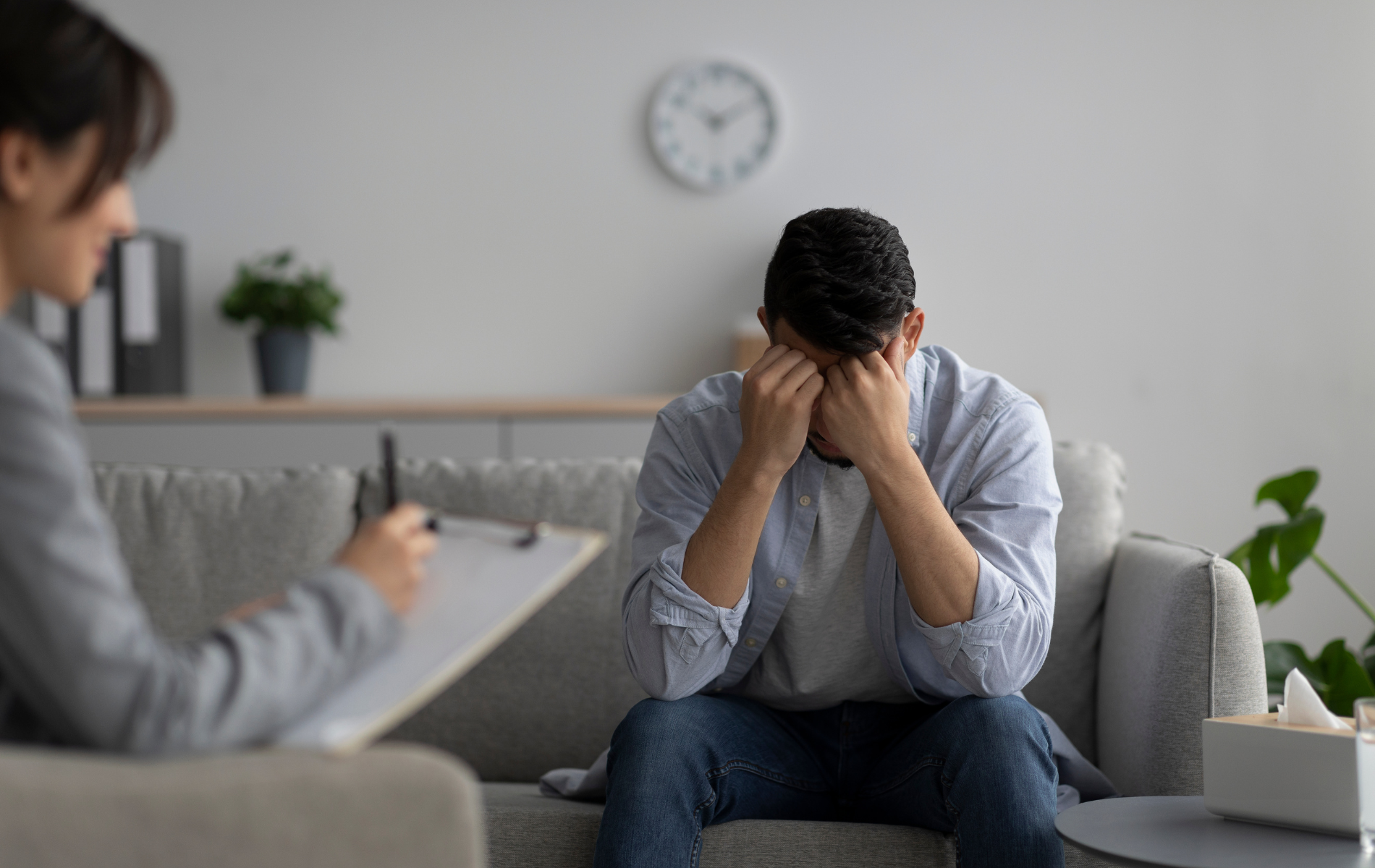 A man is sitting on a couch with his head in his hands while a woman writes on a clipboard.