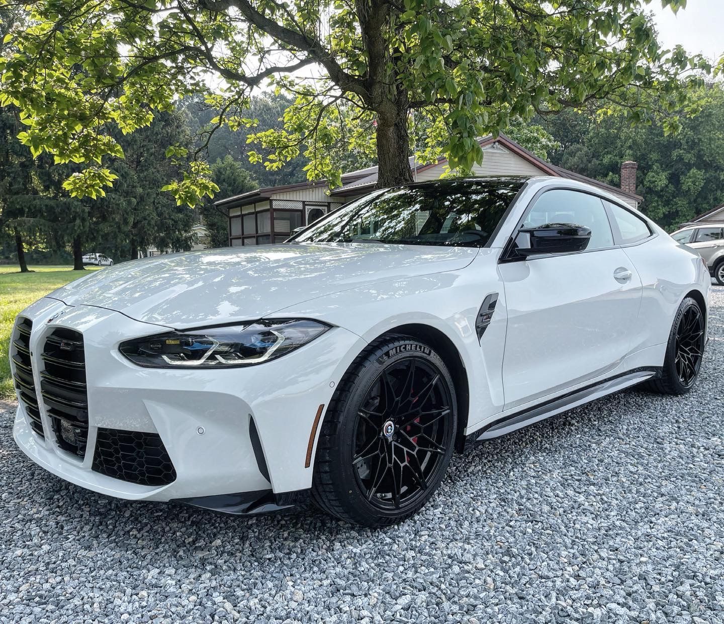 A white bmw m4 coupe is parked on gravel in front of a tree.