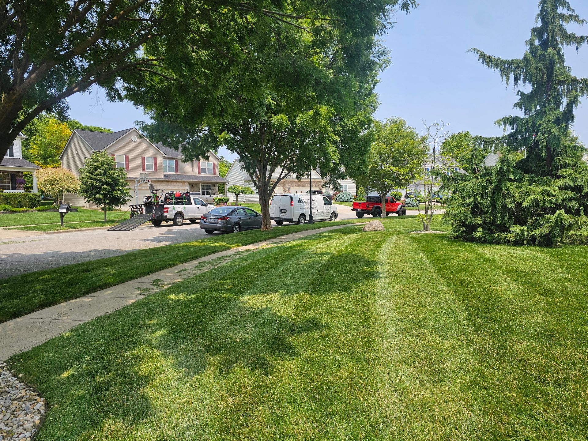 Lush green lawn with freshly cut stripes, sidewalk, street with parked vehicles, and houses under a sunny sky.
