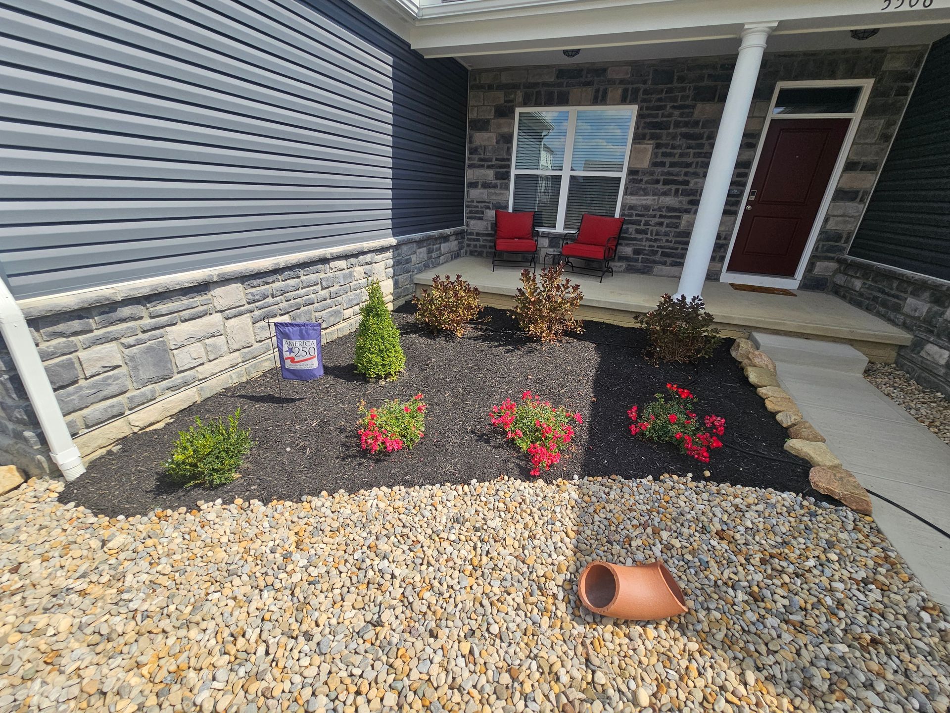 A home's front yard with dark mulch flower beds, shrubs, and red flowers. Beige gravel borders the beds.