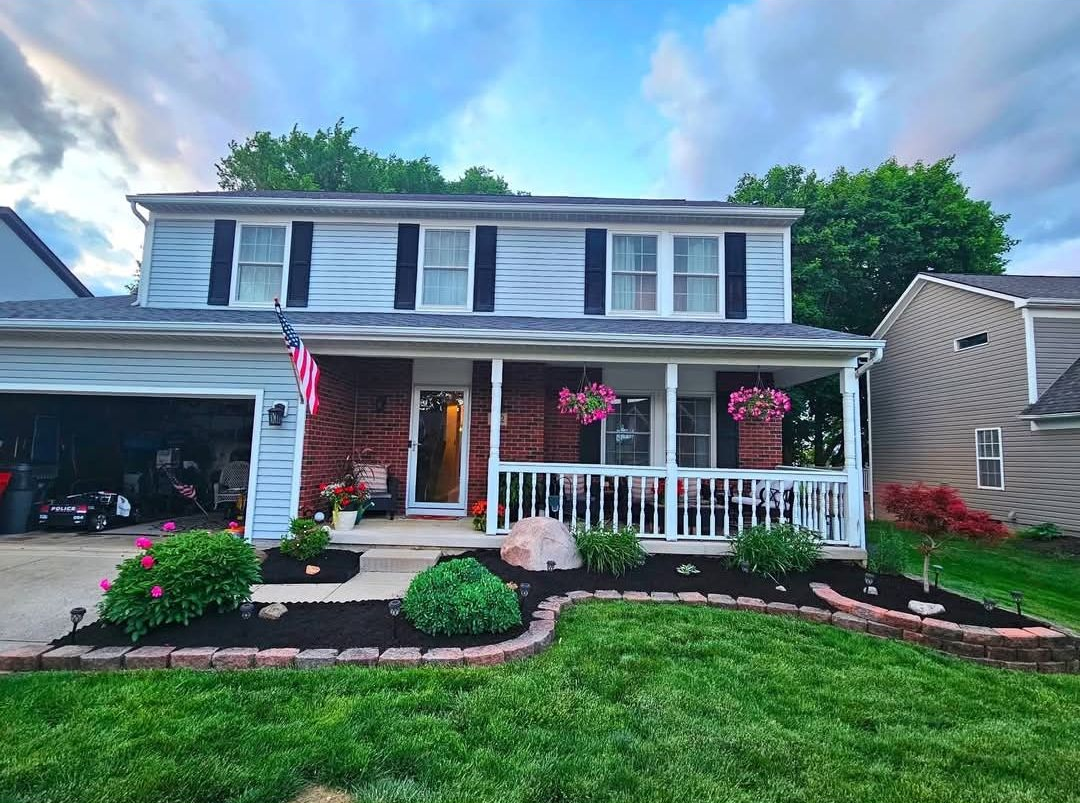 Two-story house with a brick porch and flower baskets, American flag, and landscaped front yard.