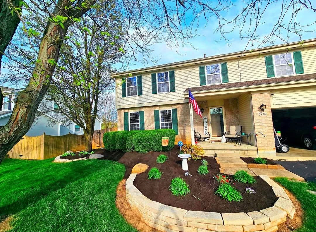 Two-story beige house with green shutters and a landscaped front yard under a blue sky.