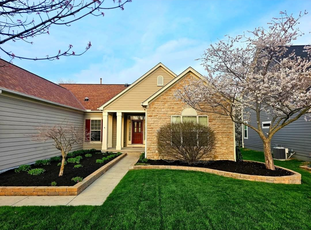 A tan and brick-fronted house with a green lawn and flowering trees.