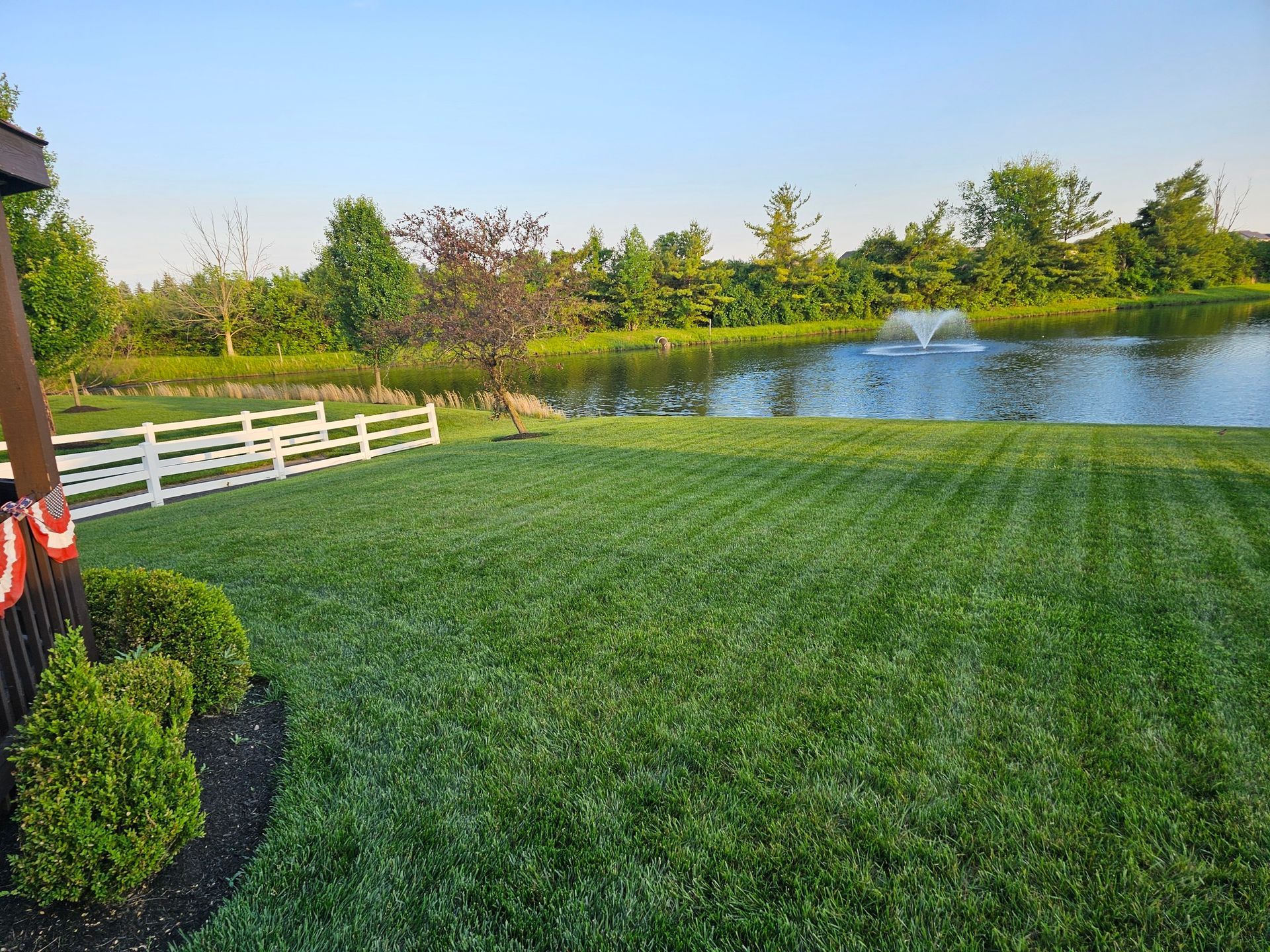 Lush green lawn leads to a pond with a fountain, white fence, trees, and a blue sky.