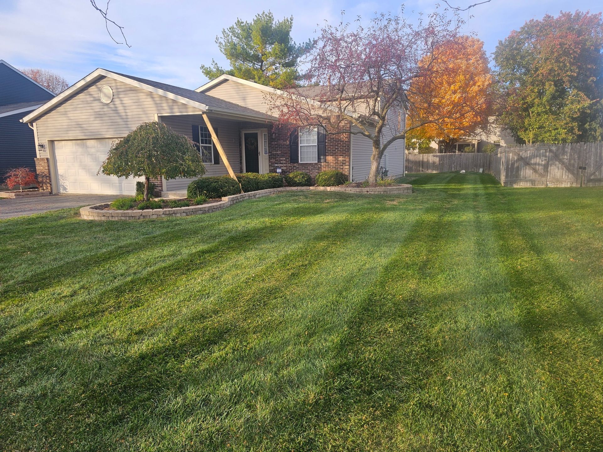 Well-manicured lawn in front of a one-story house with a garage; trees with fall foliage in the background.