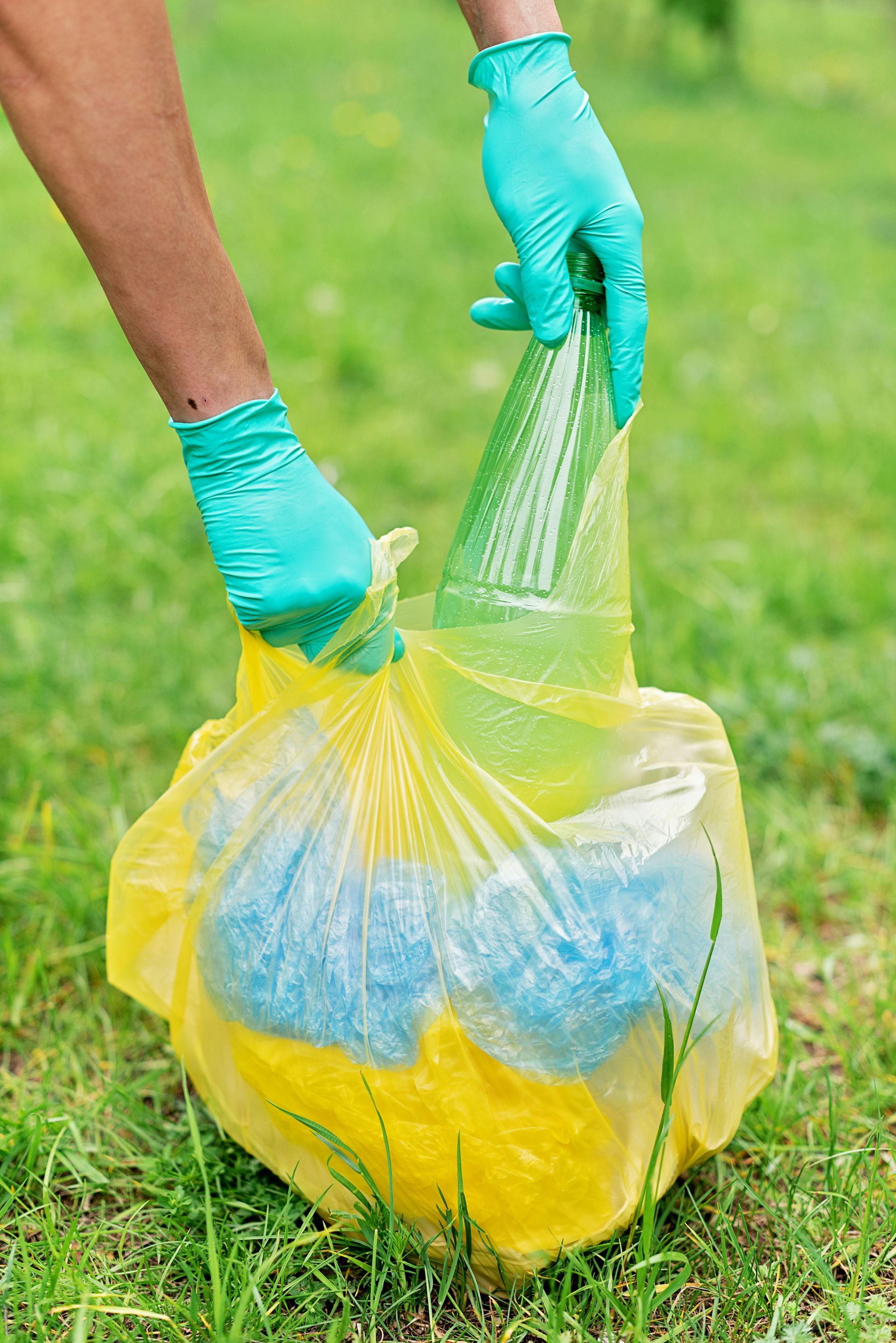 Person in teal gloves holding a yellow trash bag in a grassy area.