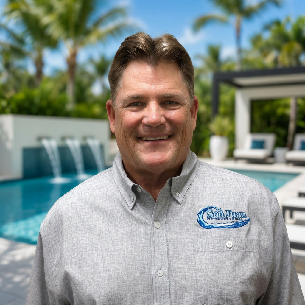 Smiling man in a gray shirt standing by a backyard pool with palm trees and patio seating.