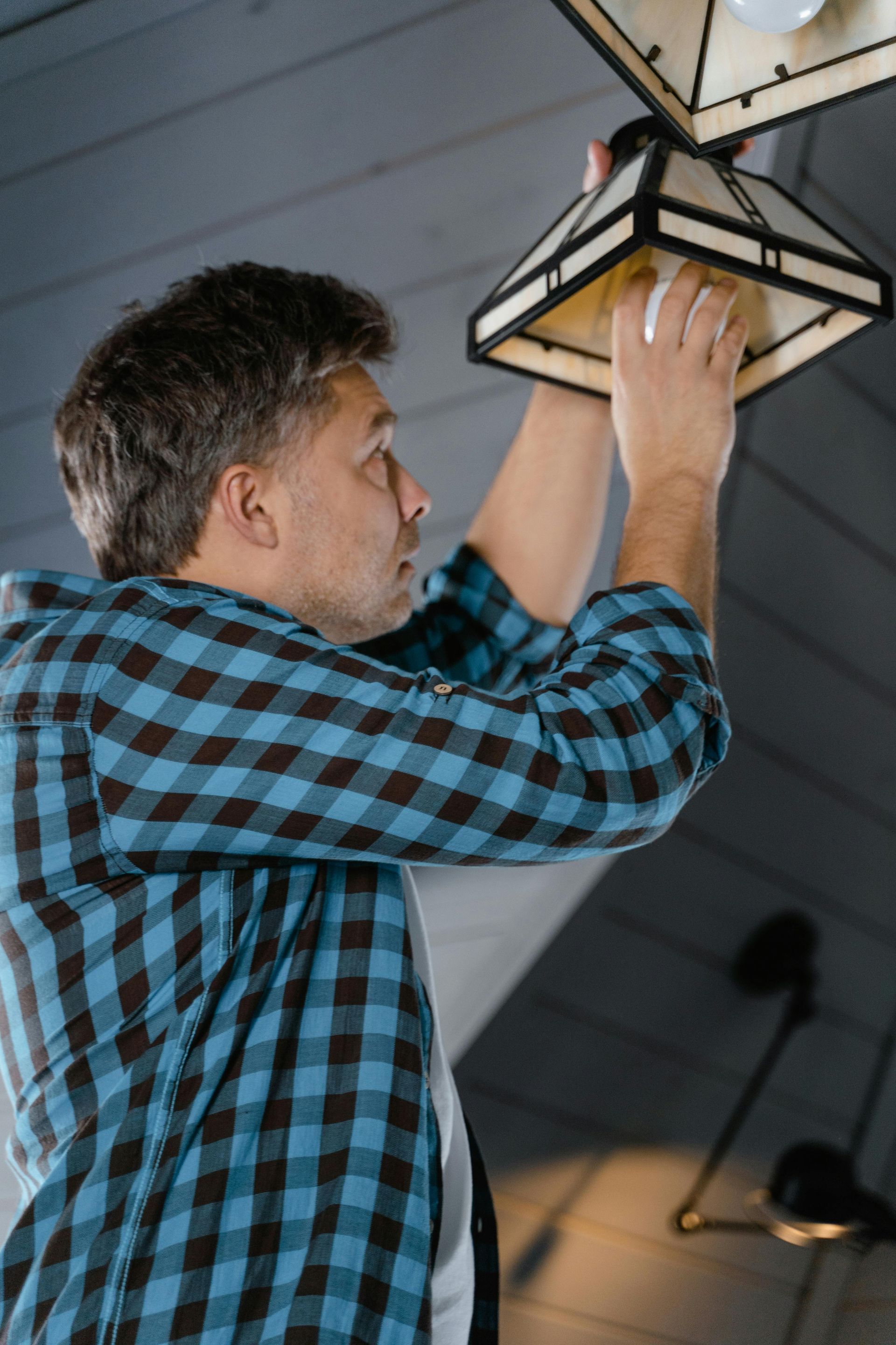 Person replacing a lightbulb in a lampshade. He wears a blue plaid shirt, in a room with a light gray ceiling.