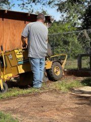 A person in a gray shirt and blue jeans operates a yellow stump grinder outdoors next to a wooden structure.