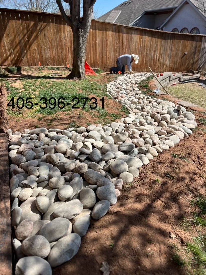 A person works on a winding dry creek bed made of large grey stones in a residential backyard.