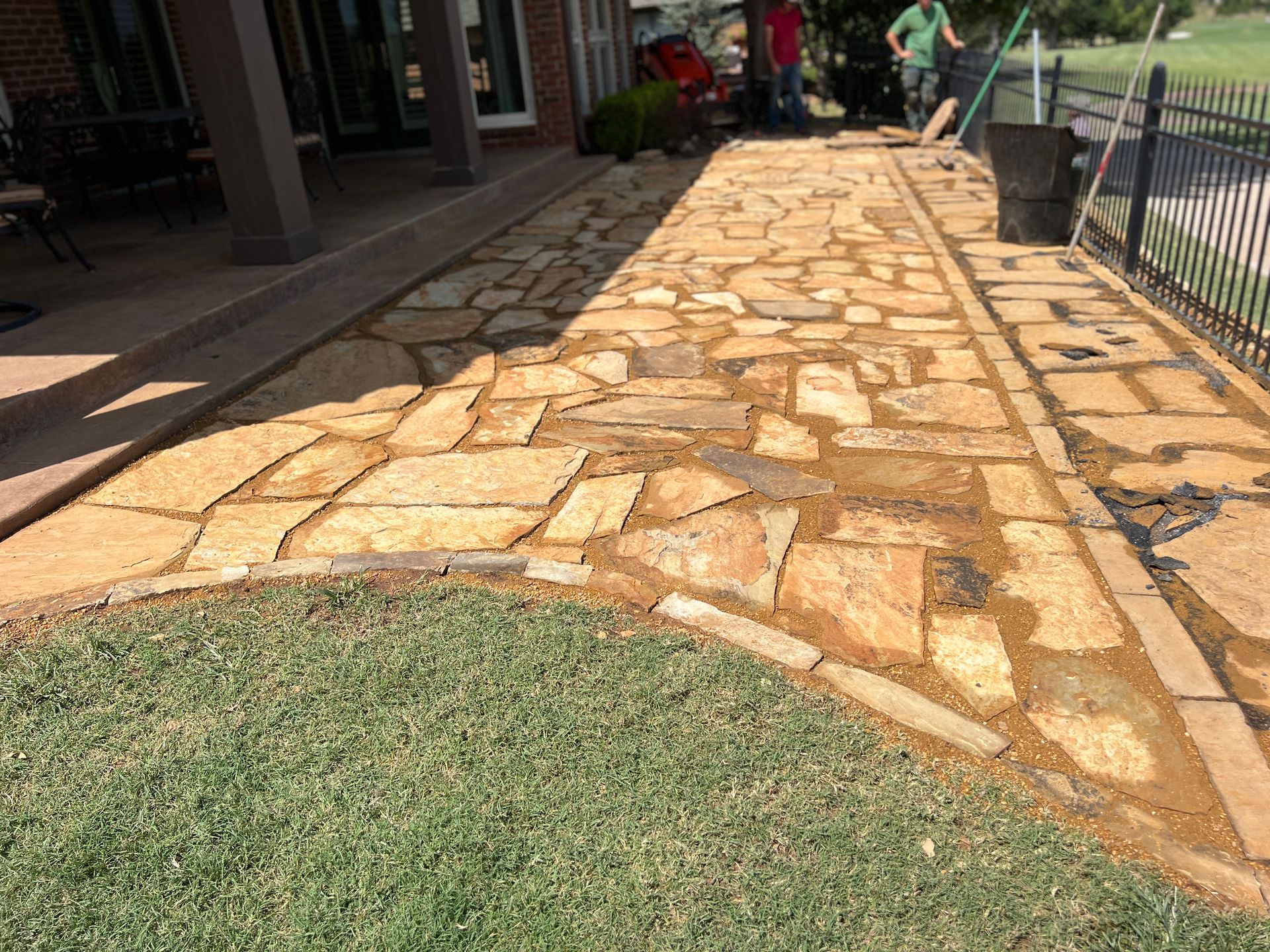 A new stone walkway sits between a grassy lawn and a patio, with people standing in the background.